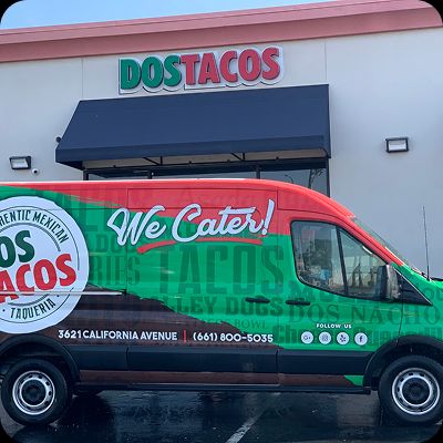 Exterior of a Dos Tacos Taqueria restaurant location with a branded black catering van parked in front. The van is decorated in red and green with 'We Cater!' and a full menu list, promoting franchise growth.