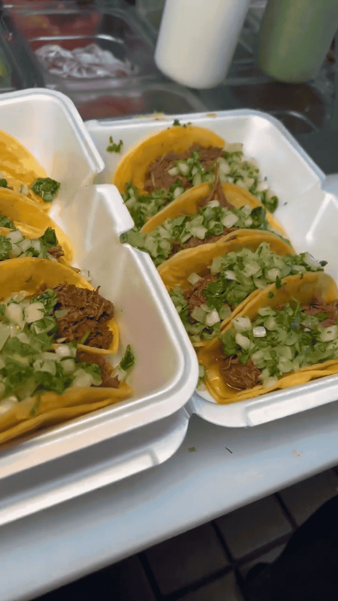 Close-up of eight street tacos in styrofoam containers, featuring beef asada, yellow corn tortillas, and fresh cilantro and onions