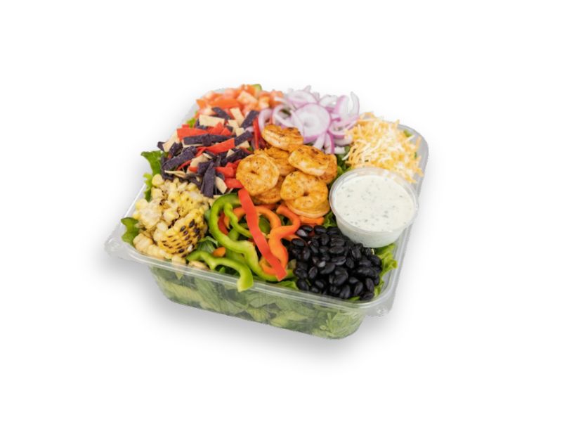A large clear plastic container of a colorful shrimp taco salad topped with grilled shrimp, black beans, grilled corn, bell peppers, tortilla strips, shredded cheese, and a side of creamy ranch dressing.