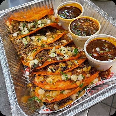Close-up of a catering aluminum tray filled with birria tacos, topped with diced onion and cilantro, served with three cups of rich, dark birria consommé for dipping.