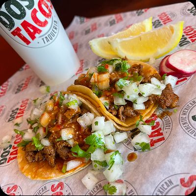Close-up of two authentic street tacos with carne asada and fresh salsa, cilantro, and onions on branded paper, next to a branded drink cup and lemon wedges.