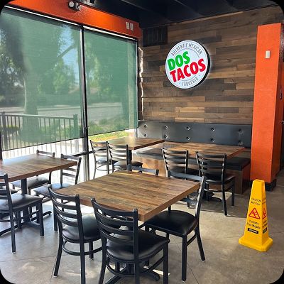 Interior dining area of the Dos Tacos Taqueria restaurant with rustic wooden tables, black chairs, a leather booth, wood-paneled walls, and the circular lit Dos Tacos logo sign.
