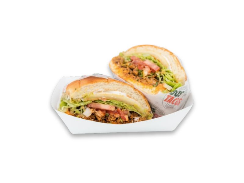 Two halves of a large Mexican Torta sandwich served in a paper tray, filled with meat, lettuce, tomato, cheese, and a sauce, wrapped partially in Dos Tacos branded paper.
